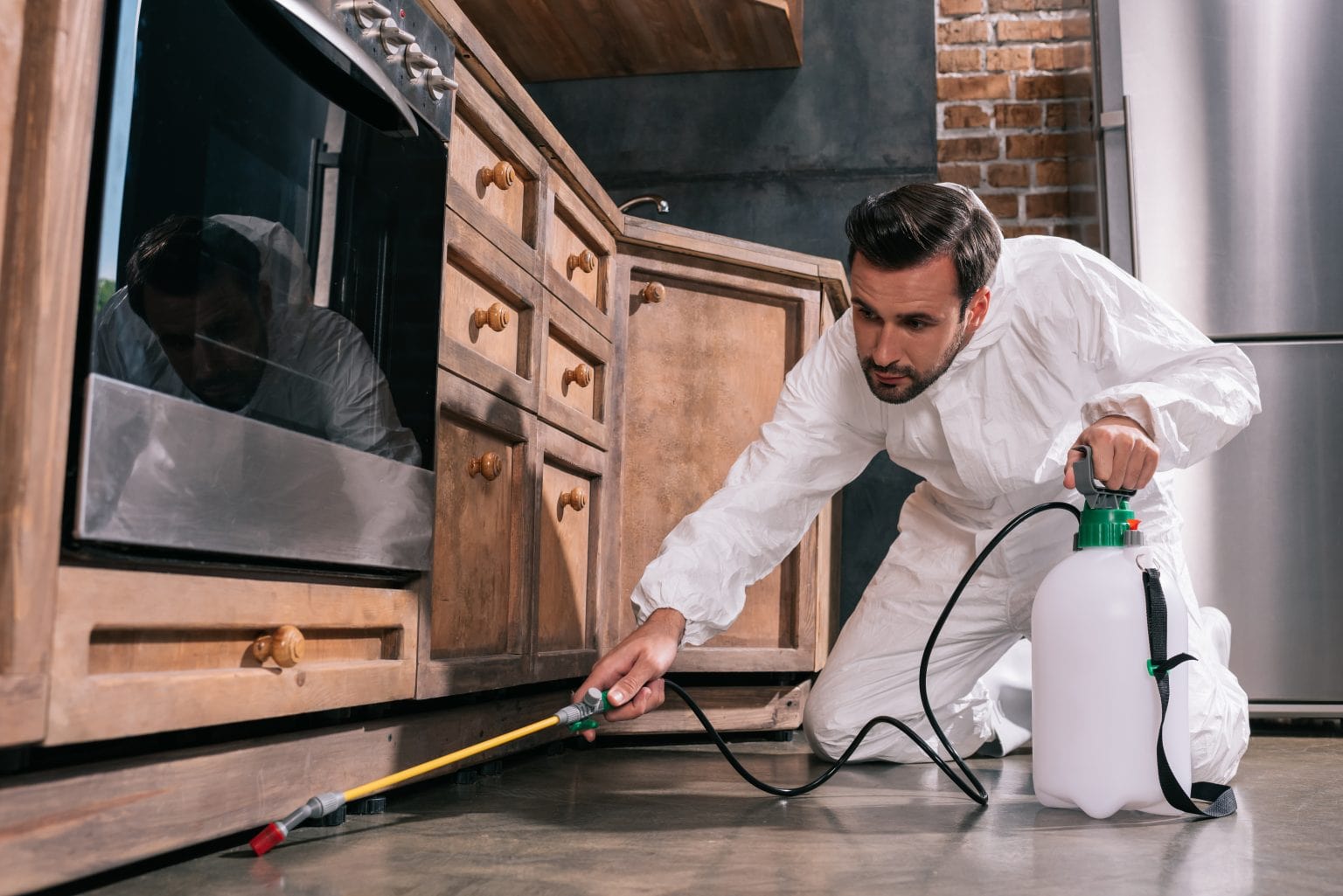Pest control technician in protective suit applying treatment under kitchen cabinets.